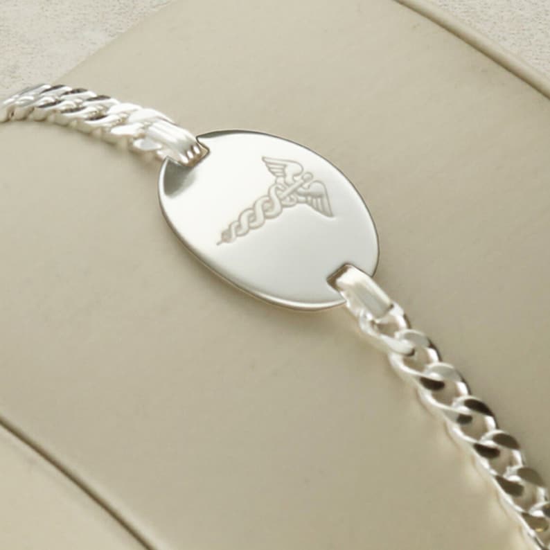 Puede incluir: Un brazalete de identificaci&oacute;n m&eacute;dica de plata con un dise&ntilde;o de cadena de eslabones y una etiqueta ovalada redonda grabada con el s&iacute;mbolo m&eacute;dico, el bast&oacute;n de Asclepio.
