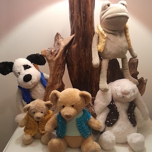 May include: A group of cuddly toys wearing knitted waistcoats. A white dog with a blue waistcoat, a brown bear with a blue waistcoat, a brown teddy bear with a blue waistcoat, a white rabbit with a black and white waistcoat, and a green frog with a yellow waistcoat.