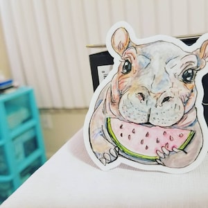 May include: A cartoon illustration of a gray hippopotamus eating a slice of watermelon. The hippopotamus has a pink nose and large eyes. The watermelon is pink with black seeds.