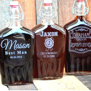 May include: Three glass bottles with personalized engravings. The bottles are brown and have a metal hinged lid. The engravings include the names "Mason", "Jaxon", and "Graham" with titles such as "Best Man", "Groomsman", and "Groom".