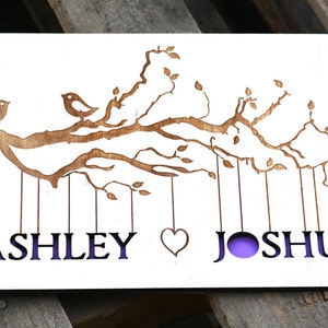 May include: A wooden guest book with a laser-cut design of birds on a tree branch. The names "ASHLEY" and "JOSHUA" are cut out below the branch, with a heart symbol between them. The background is a light purple color.