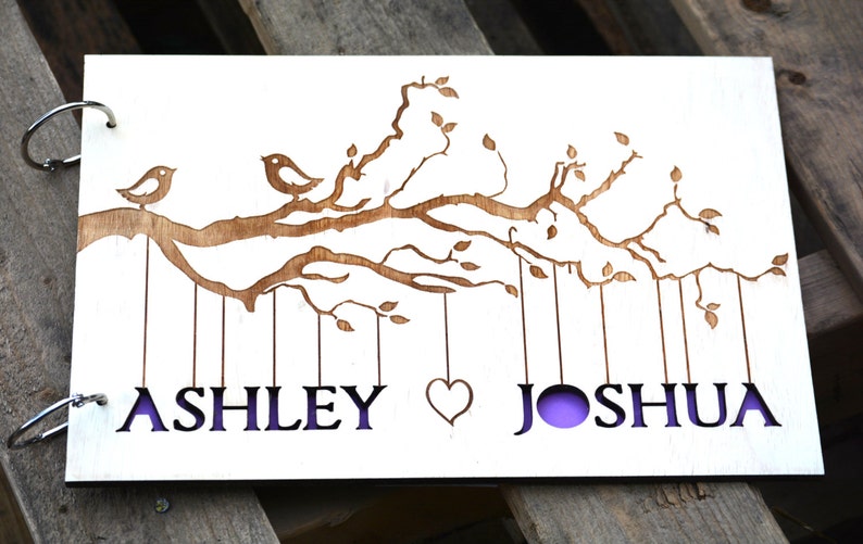 May include: A wooden guest book with a laser-cut design of birds on a tree branch. The names "ASHLEY" and "JOSHUA" are cut out below the branch, with a heart symbol between them. The background is a light purple color.
