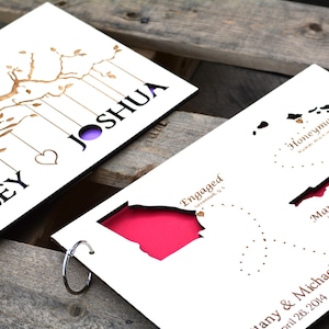 May include: A white paper wedding keepsake with laser-cut designs. One side features a tree with the names "Ashley" and "Joshua." The other side shows states with the words "Engaged," "Honeymoon," and "Married." The background is a rustic wooden surface.