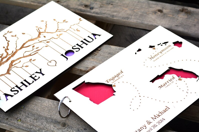 May include: A white paper wedding keepsake with laser-cut designs. One side features a tree with the names "Ashley" and "Joshua." The other side shows states with the words "Engaged," "Honeymoon," and "Married." The background is a rustic wooden surface.