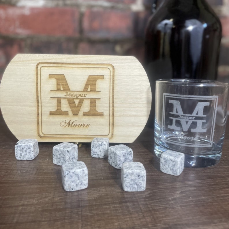May include: A wooden tray with a personalised engraved design featuring the initials "M" and the name "Jasper Moore". The tray is accompanied by a glass tumbler with the same engraved design and a set of eight grey granite whiskey stones.