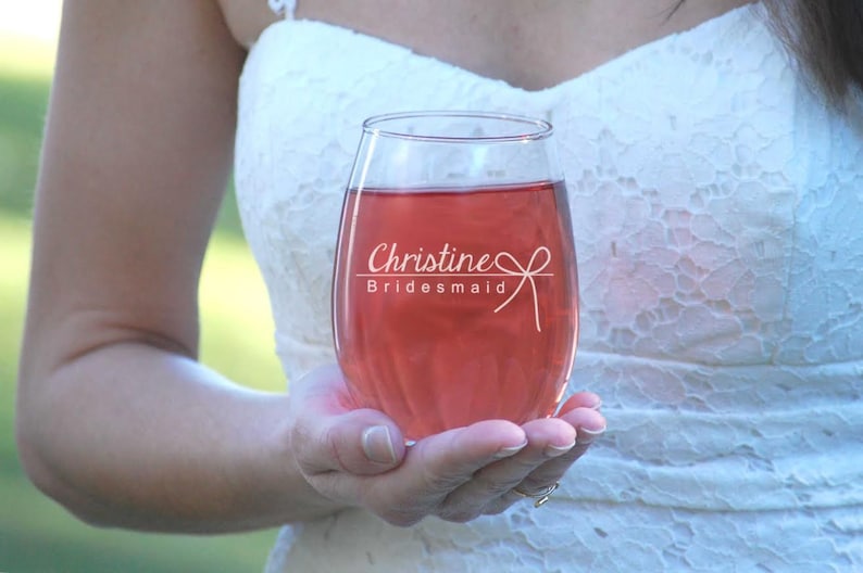 May include: A clear stemless wine glass with the text "Christine Bridesmaid" etched on the side. The glass is filled with a pink beverage.