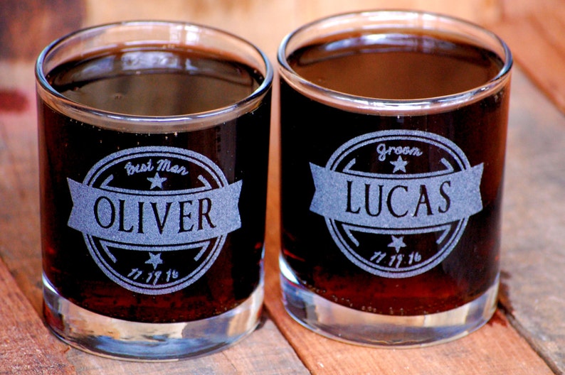 May include: Two clear glass tumblers filled with a dark liquid. The tumblers are engraved with a circular design and the text "Best Man OLIVER" and "Groom LUCAS".