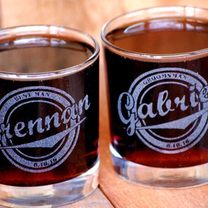 May include: Two clear glass tumblers filled with a dark liquid. The glasses are engraved with a circular design and the text "Best Man Brennan 6.10.16" and "Groomsman Gabriel 6.10.16".