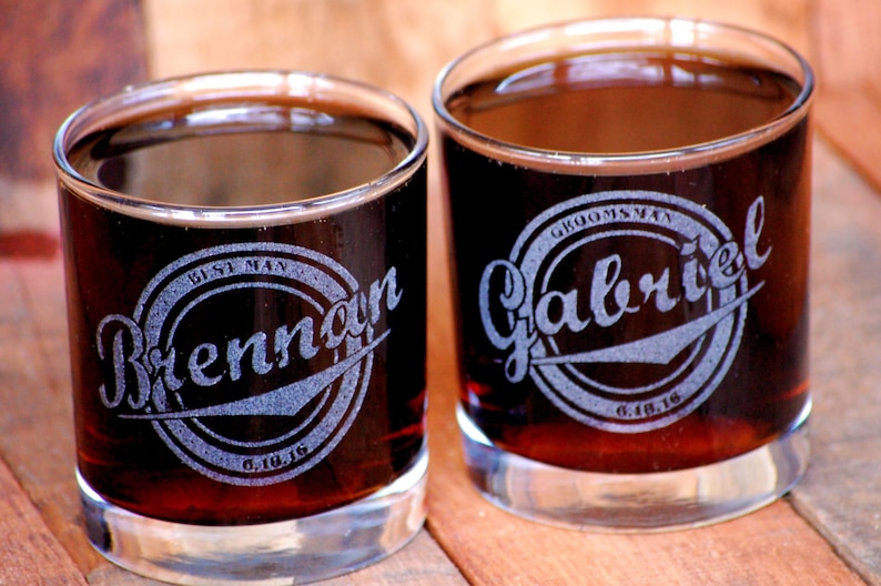 May include: Two clear glass tumblers filled with a dark liquid. The glasses are engraved with a circular design and the text "Best Man Brennan 6.10.16" and "Groomsman Gabriel 6.10.16".