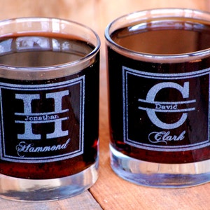 May include: Two clear glass tumblers filled with a dark liquid. Each tumbler features a personalised monogram with a name and initial etched into the glass.