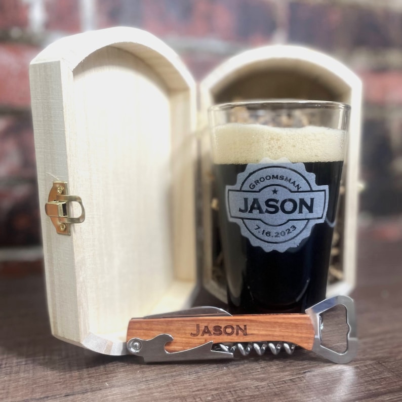 Puede incluir: Una caja de madera con tapa con bisagras, que contiene un vaso de pinta de vidrio transparente con una bebida negra y una inscripci&oacute;n personalizada que dice "Groomsman Jason 7.16.2023". Un abridor de botellas de madera con un sacacorchos y una inscripci&oacute;n personalizada que dice "Jason" est&aacute; delante de la caja.