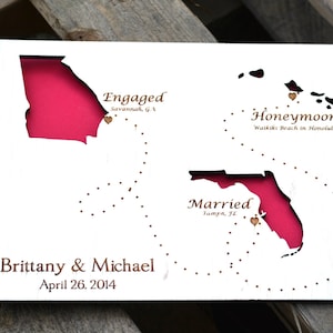 May include: A white wooden map with cutouts of U.S. states, including Georgia, Florida, and Hawaii, with a pink background. Text includes "Engaged," "Honeymoon," and "Married," along with names and a date.