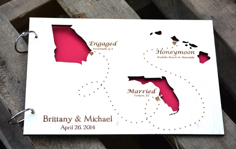 May include: A white wooden map with cutouts of U.S. states, including Georgia, Florida, and Hawaii, with a pink background. Text includes "Engaged," "Honeymoon," and "Married," along with names and a date.