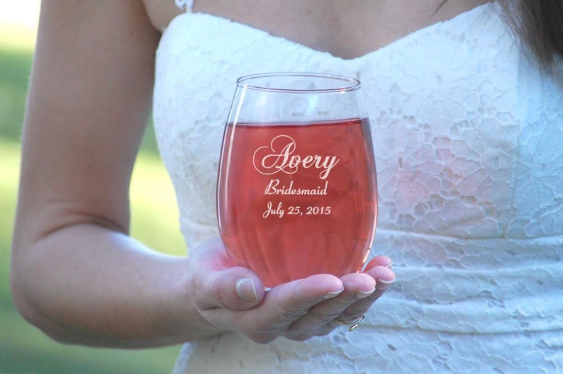 May include: A clear glass wine glass with a pink beverage inside. The glass is engraved with the text "Avery Bridesmaid July 25, 2015".