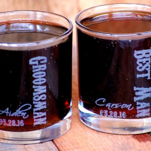 May include: Two clear glass tumblers filled with a dark liquid. One tumbler is engraved with "Best Man" and the date "05.28.16" and the other is engraved with "Groomsman" and the date "05.28.16".