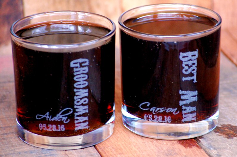 May include: Two clear glass tumblers filled with a dark liquid. One tumbler is engraved with "Best Man" and the date "05.28.16" and the other is engraved with "Groomsman" and the date "05.28.16".