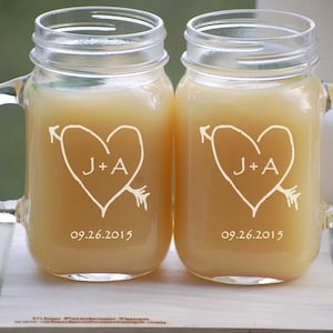 May include: Two clear glass mason jar mugs with handles. Each mug has a white heart with an arrow through it and the initials "J+A" inside the heart. The date "09.26.2015" is printed below the heart.