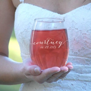 May include: A clear glass wine glass with a personalized inscription that reads "courtney 06.21.2015". The glass is filled with a pink beverage.