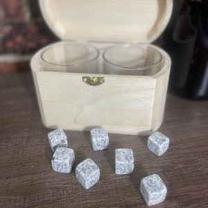 May include: A wooden box with a hinged lid, containing two clear glass tumblers and a set of grey granite whisky stones. The box is open and the stones are scattered on a wooden surface.