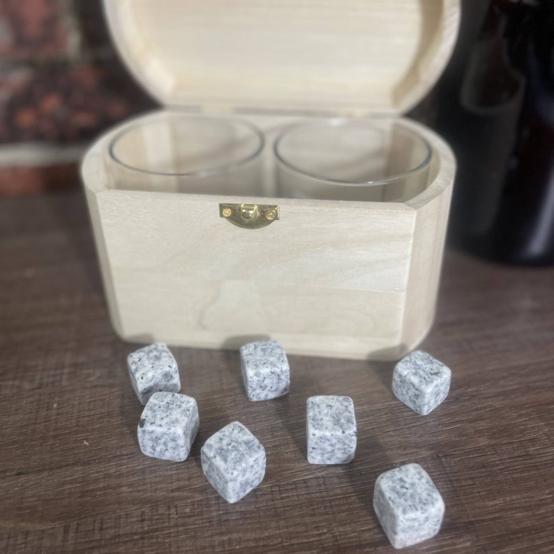 May include: A wooden box with a hinged lid, containing two clear glass tumblers and a set of grey granite whisky stones. The box is open and the stones are scattered on a wooden surface.