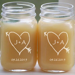 May include: Two clear glass mason jars with handles, each filled with a light brown liquid. The jars are engraved with a heart design, an arrow, and the initials "J+A" and the date "09.26.2015".