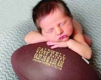 May include: A brown leather football with the name "Caldwell Wayne Rainey" and the date "04.27.16 @ 1:15 AM" engraved on it. A newborn baby is sleeping on top of the football.