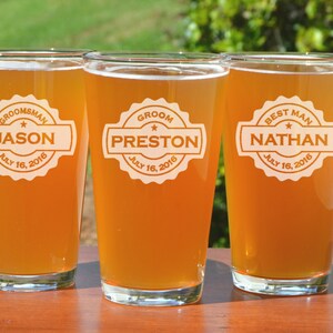 Puede incluir: Tres jarras de cerveza de vidrio transparente con etiquetas personalizadas. Las etiquetas son blancas con texto negro y una estrella. Las jarras est&aacute;n llenas de una bebida de color &aacute;mbar claro. Las etiquetas dicen "Groom, Preston, July 16, 2016", "Best Man, Nathan, July 16, 2016", y "Groomsman, Jason, July 16, 2016".