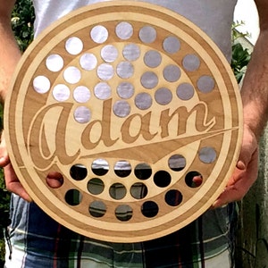 May include: A round wooden bottle opener with a brown wood grain finish. The opener has a circle of holes for bottle caps and the name "Adam" is engraved in a script font.