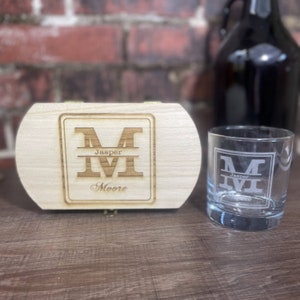 May include: A wooden box with a personalised engraved design featuring the letter "M" with the name "Jasper Moore" underneath. The box is next to a clear glass with the same engraved design.