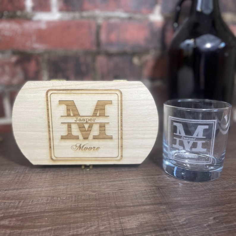 May include: A wooden box with a personalised engraved design featuring the letter "M" with the name "Jasper Moore" underneath. The box is next to a clear glass with the same engraved design.