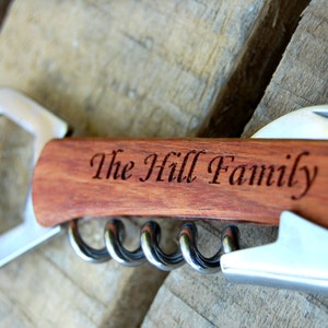 May include: A wooden bottle opener with a corkscrew and a bottle opener. The handle is engraved with the text "The Hill Family".