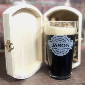 Puede incluir: Un vaso de pinta personalizado con el texto "Groomsman Jason 7.16.2023" en un c&iacute;rculo negro con una estrella. El vaso est&aacute; en una caja de madera con un cierre.