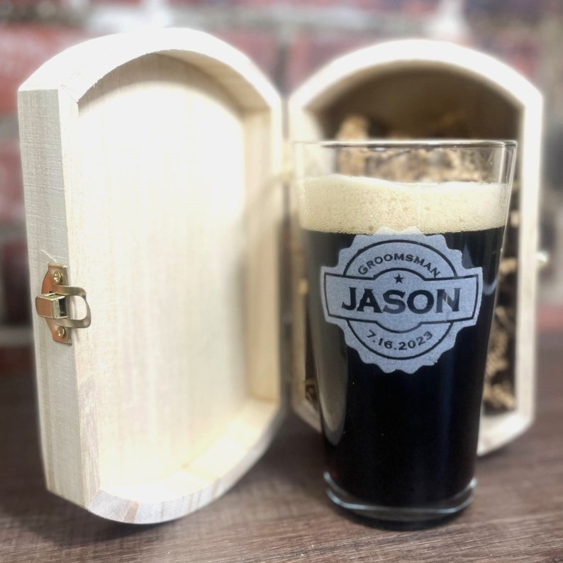 Puede incluir: Un vaso de pinta personalizado con el texto "Groomsman Jason 7.16.2023" en un c&iacute;rculo negro con una estrella. El vaso est&aacute; en una caja de madera con un cierre.