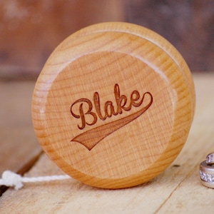 May include: A wooden yo-yo with the name "Blake" engraved on it. The yo-yo is light brown and has a string attached to it. There are two silver rings with diamonds on them in the background.