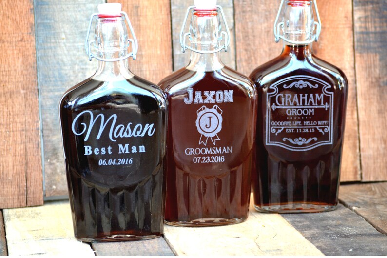 Op de afbeelding: Drie glazen flessen met gepersonaliseerde gravures. De flessen zijn bruin en hebben een metalen scharnierdeksel. De gravures omvatten de namen "Mason", "Jaxon" en "Graham" met titels zoals "Best Man", "Groomsman" en "Groom".