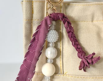 May include: A decorative keychain charm with a gold-colored clasp. The charm features a braided dark purple fabric accent, a beaded strand with white and clear beads, and a dark purple fabric accent with white stitching. The charm is attached to a beige tote bag.