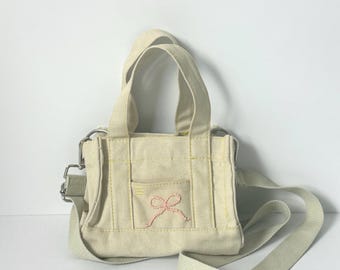May include: A small, cream-colored canvas tote bag with a pink embroidered bow on the front pocket. The bag has a matching adjustable shoulder strap and two short handles. Yellow stitching accents the seams.
