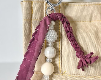 May include: A bag charm featuring a silver-toned metal lobster clasp, a braided strip of dark maroon fabric with white stitching, and a vertical strand of beads including clear faceted spheres, a smooth white bead, and a cream-colored bead.