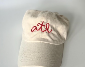 May include: A cream-colored baseball cap featuring the lowercase letters "atl" embroidered in red thread on the front panel. The cap has a curved brim, a six-panel construction with stitched eyelets, and a structured crown design.