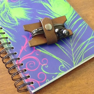 May include: A spiral-bound notebook with a purple cover featuring green and pink feather-like designs. A brown leather earbud holder with earbuds is on the notebook. A blue and clear pen rests on a wooden surface.