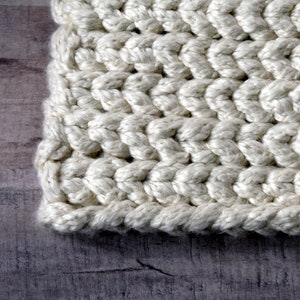 May include: Close-up of a section of a cream-colored, chunky knit fabric. The thick yarn is tightly woven, creating a textured surface with visible stitches. The fabric's edges are neatly finished, suggesting a handmade item, possibly a blanket or scarf.