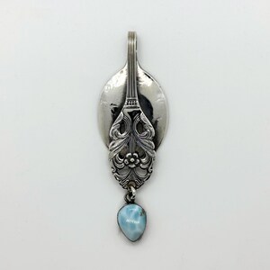 May include: A silver pendant crafted from a spoon, featuring ornate floral detailing and a light blue, teardrop-shaped gemstone. The pendant has a loop at the top for a chain. The gemstone is approximately 0.75 inches in length.