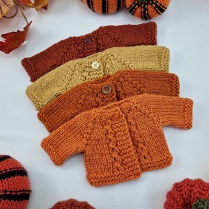 May include: Four hand-knitted baby cardigans in shades of orange, rust, and gold are displayed. The cardigans are arranged in a row, each with a button closure. Decorative pumpkins and autumn leaves surround the cardigans, creating a seasonal theme.