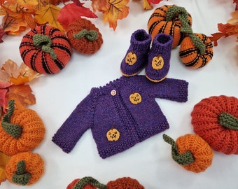 May include: A purple knitted long-sleeve cardigan with a single button closure and two small crocheted jack-o'-lantern appliques, accompanied by a pair of matching purple knitted booties, each featuring a small crocheted jack-o'-lantern applique.