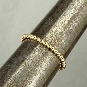 May include: A delicate gold ring featuring a beaded design. The ring is composed of small, evenly spaced golden beads, creating a textured and elegant appearance. The ring is shown on a silver metal surface.