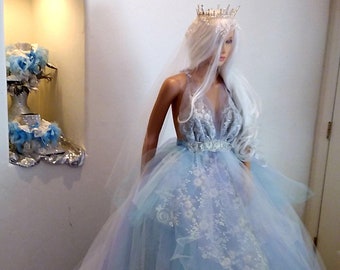 May include: A pale blue and lilac tulle wedding dress on a mannequin, with a low neckline and floral embroidery. The dress has a long train and is accessorized with a silver crown and veil. The gown is displayed in a well-lit room.
