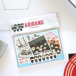 May include: A sticker pack labeled "ARIRANG" with various cartoon illustrations of a group of people. The stickers feature images of a ship, an alien spacecraft, and the words "BODY TO BODY" and "HANN HOCAG". The pack is on a light blue background.