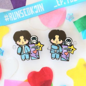 May include: Two enamel pins featuring a cartoon character in a light blue outfit, holding a microphone and a sign with a heart and star. The pins are surrounded by colorful confetti and a light blue ribbon with text.