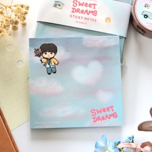 May include: A square notepad with a light blue background featuring a cartoon character holding flowers, floating in a sky with pink clouds. The words "Sweet Dreams" are in pink. The notepad is part of a stationery set.