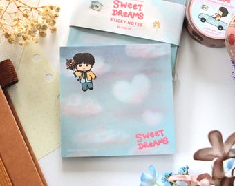 May include: A square notepad with a light blue background featuring a cartoon character holding flowers, floating in a sky with pink clouds. The words "Sweet Dreams" are in pink. The notepad is part of a stationery set.